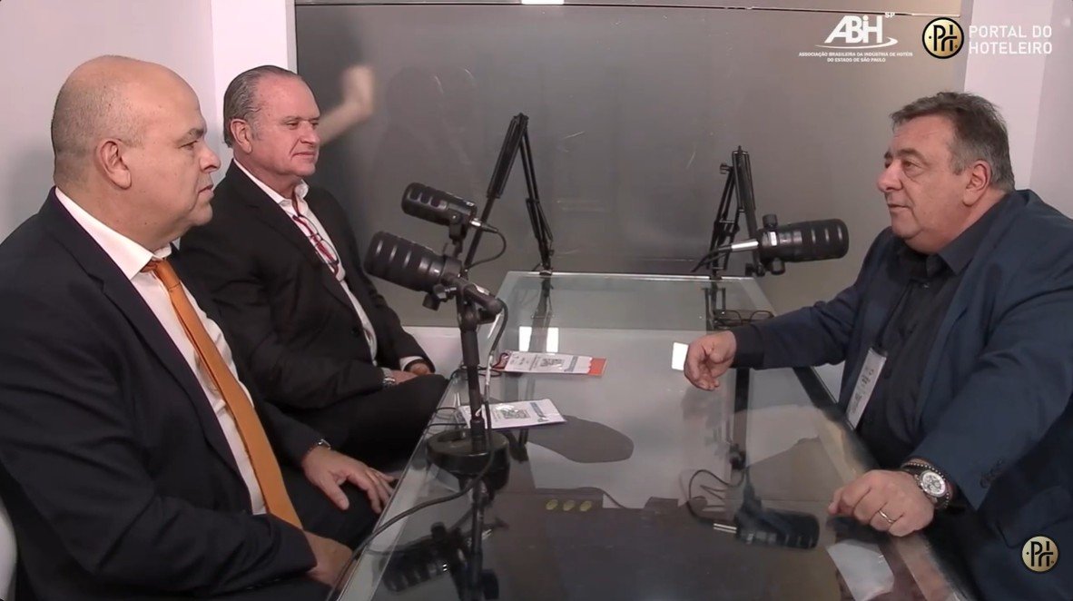 Três homens em trajes formais participam de uma entrevista em um estúdio de videocast. À direita, Luiz Henrique Miranda (Portal do Hoteleiro) conduz a conversa com Anselmo Santana (TOLON) e João Ebert (Sill do Brasil), sentados à esquerda. Eles estão ao redor de uma mesa de vidro com microfones profissionais. No canto superior, aparecem os logotipos da ABIH-SP e do Portal do Hoteleiro. A imagem ilustra um debate sobre inovação tecnológica na lavanderia hoteleira.