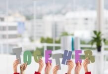 Representatividade que gera resultado: por que sou adepto do associativismo Fotografia conceitual que ilustra o associativismo através de várias mãos unidas erguendo letras coloridas que formam a palavra "TOGETHER" (juntos). Cada letra possui uma cor e textura diferente, simbolizando a união de diversos indivíduos em prol de um objetivo comum. O fundo é um ambiente de escritório claro e moderno, reforçando a ideia de colaboração profissional.