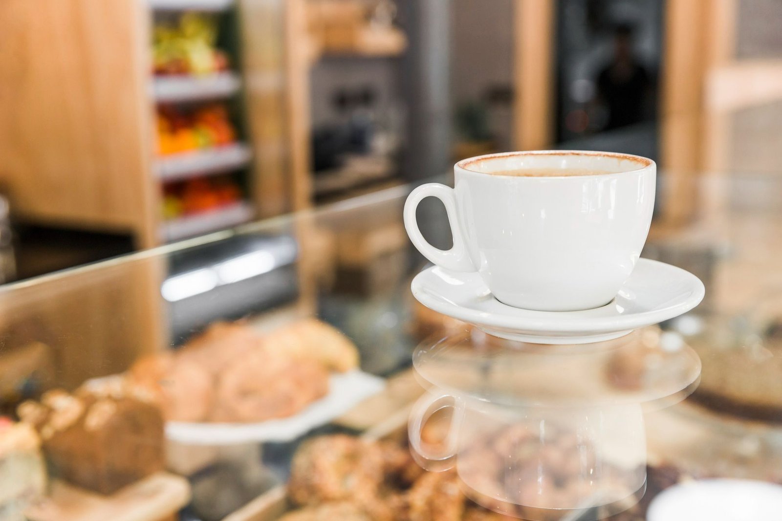 Uma xícara de café branca sobre uma mesa de vidro em um lobby de hotel. Ao fundo, de forma suavemente desfocada, vê-se uma vitrine iluminada de cafeteria com opções de bolos, pães e doces. A imagem destaca a conveniência do serviço de A&B (Alimentos e Bebidas) como uma oferta de valor e conforto para hóspedes e visitantes no ambiente de recepção.