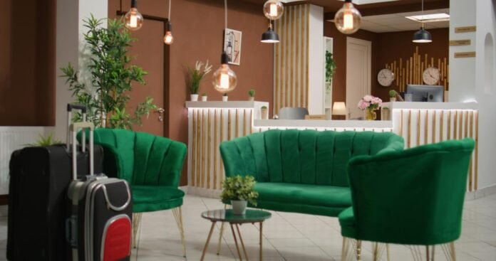 Um aconchegante lobby de hotel com paredes em tom de marrom quente e iluminação pendente suave. Destaque para um conjunto de sofás e poltronas de veludo verde-esmeralda posicionados ao redor de uma pequena mesa de centro. Ao fundo vê-se o balcão da recepção iluminado com detalhes em madeira, plantas decorativas e malas de viagem em primeiro plano, transmitindo uma atmosfera de hospitalidade e conforto.