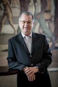Retrato de Orlando de Souza, presidente do FOHB, um homem de pele clara, cabelos grisalhos e óculos de armação clara. Ele sorri levemente para a câmera, veste um paletó escuro sobre uma camisa social listrada e está com as mãos entrelaçadas à frente do corpo. Ao fundo, há uma pintura artística em tons terrosos levemente desfocada.