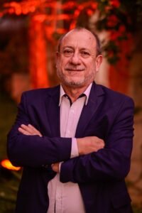 Retrato profissional de Toni Sando, presidente da Unedestinos e do Visite São Paulo, em plano médio. Ele aparece com os braços cruzados, sorrindo diretamente para a câmera, e veste um blazer azul-marinho sobre uma camisa social de cor clara. O fundo é composto por folhagens envoltas em uma iluminação quente e avermelhada, o que sugere um ambiente de evento noturno ou confraternização.