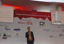 Troféu Fornecedor Destaque da Hotelaria merece muitos aplausos Edgar Oliveira, diretor editorial da Revista Hotéis, em pé no centro de um palco durante a abertura do Troféu Fornecedor Destaque da Hotelaria 2025. Ele veste blazer escuro, camiseta bege e tênis brancos, segurando um microfone enquanto fala. Ao fundo, um painel branco e vermelho exibe o nome do evento e logotipos de parceiros como FBHA, Resorts Brasil, Sindal, R1 Audiovisual, Wyndham e outros. À esquerda, uma tela projeta o nome do troféu e, à direita, outra tela mostra a imagem da estatueta da premiação.