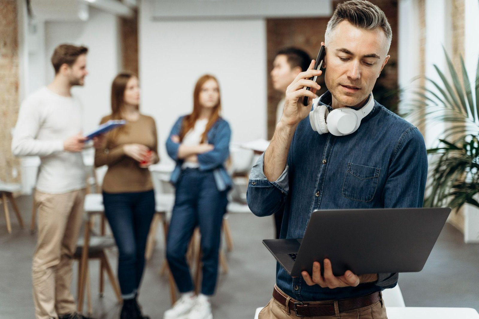 Um homem grisalho, com fones de ouvido no pescoço, de pé em um escritório aberto, foca em um notebook aberto enquanto fala ao celular com uma expressão atenta. Ao fundo, borrados, outros colaboradores conversam. A imagem ilustra a ação de receber informações e agir de forma proativa, exemplificando a importância de dar feedback e tomar providências após utilizar canais de escuta na empresa.