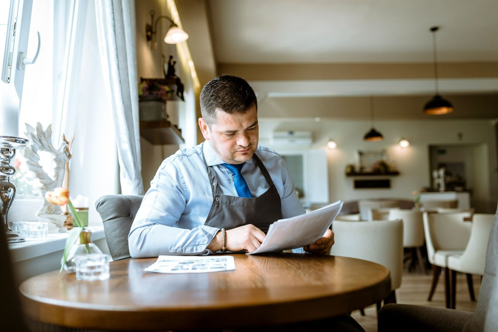 Homem de camisa social azul, gravata e avental preto, sentado à mesa de um restaurante, analisa com atenção um material impresso. A foto pretende ilustrar a importância da atualização profissional para o êxito do estabelecimento. 