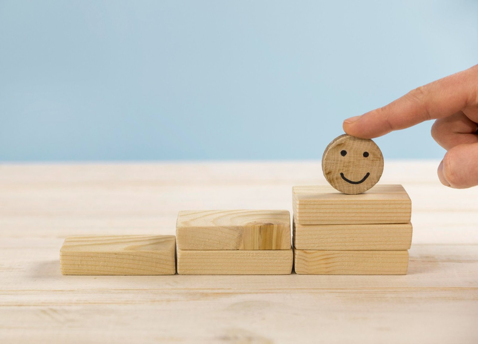 Uma mão humana toca um disco de madeira com um rosto sorridente, posicionado no topo de uma escada feita de blocos retangulares. A cena, sobre uma mesa de madeira clara e fundo azul, representa a conquista de um objetivo e a satisfação profissional.