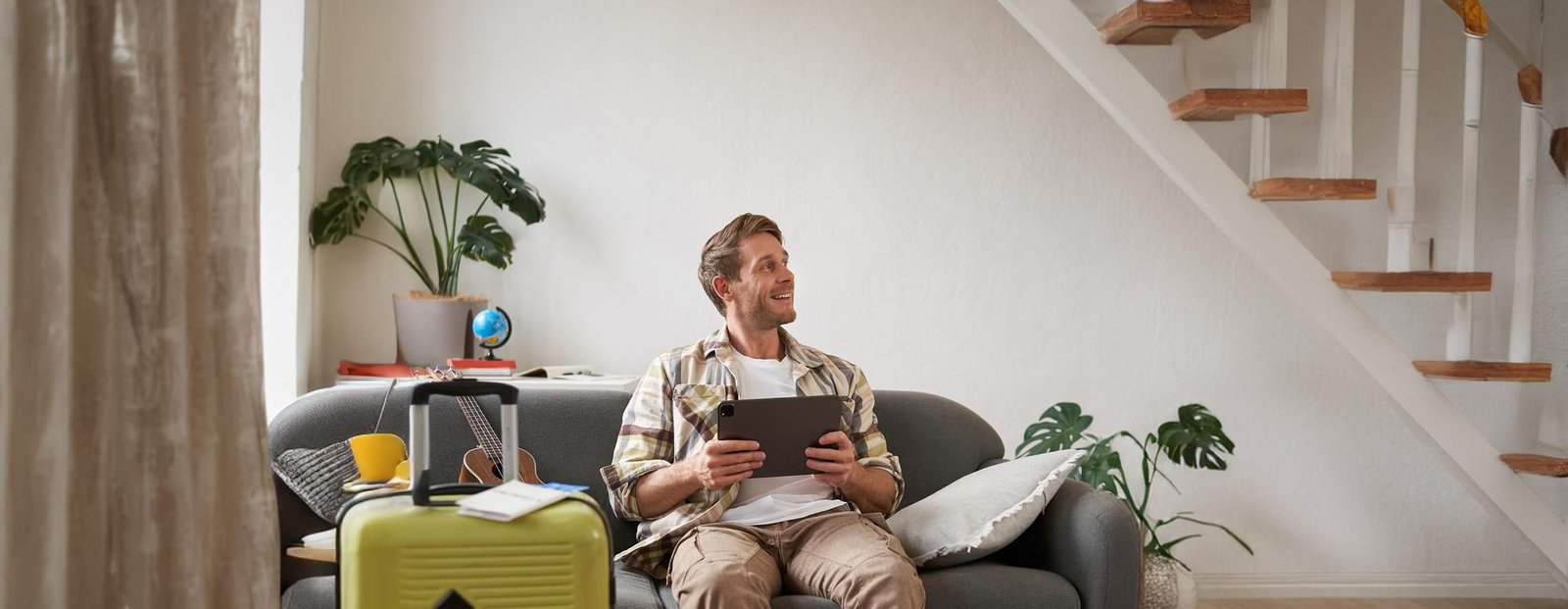 Um homem sorridente sentado em um sofá cinza, segurando um tablet e olhando para cima, em uma expressão contemplativa. À sua frente, uma mala verde com tickets de passagem em cima sugere que ele está prestes a viajar. O ambiente é iluminado e sugere o momento de planejamento ou de antecipação da viagem.