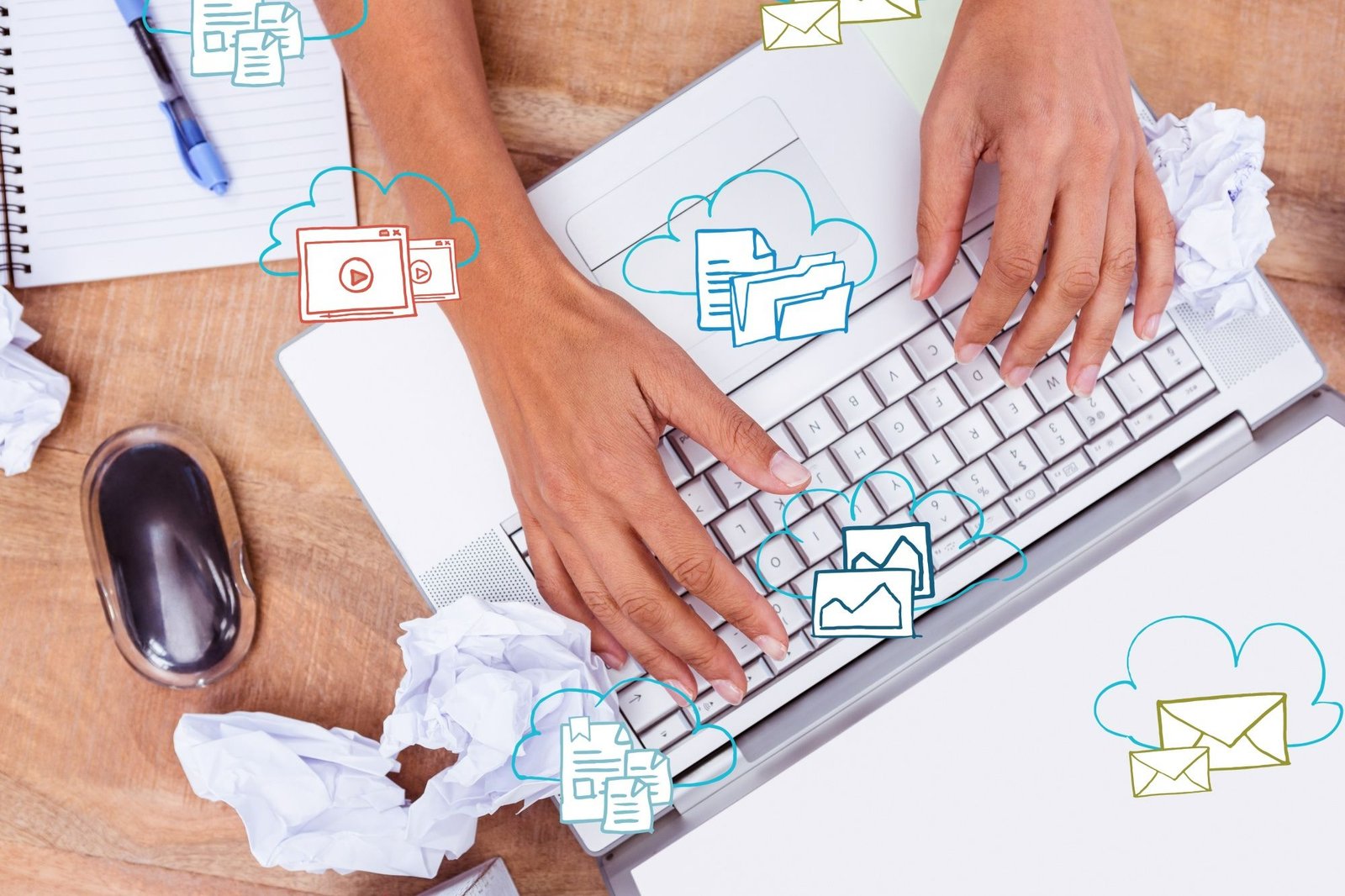 Vista superior de mãos digitando em um laptop sobre uma mesa de madeira. Ao redor do computador, há ilustrações digitais de nuvens contendo ícones de envelopes, vídeos e arquivos, simbolizando a organização de uma estratégia de e-mail marketing para hotéis. Bolas de papel amassado e um caderno ao lado sugerem o processo criativo de elaboração das campanhas.