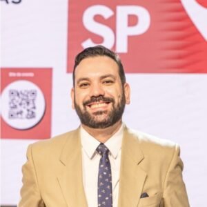 Retrato do Fabio Zelenski, um homem de barba e bigode, sorrindo e vestindo um paletó bege sobre uma camisa branca e gravata estampada. Ele está posicionado em frente a um fundo abstrato com um grande "SP" e um código QR visível à esquerda.
