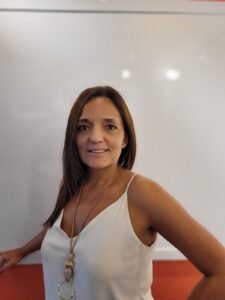 Retrato de meio corpo da executiva Lucila Quintino, fundadora e líder da HotelConsult, sorrindo para a câmera. Ela é uma mulher de cabelos castanhos longos e lisos, veste uma regata branca de alças finas e um colar longo artesanal com pingentes redondos. O fundo é minimalista e desfocado, combinando uma parede branca e uma superfície laranja na parte inferior.