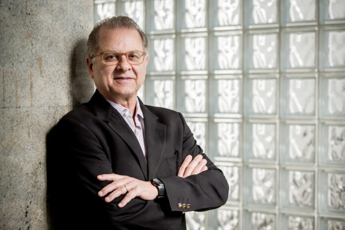 Retrato corporativo de Orlando de Souza, presidente do FOHB e autor deste artigo sobre canais de distribuição na hotelaria. Homem de meia-idade, com cabelos grisalhos e óculos de grau, veste terno preto sobre camisa de listras finas e posa com braços os cruzados. Ele sorri para a câmera e usa um relógio de pulso preto.