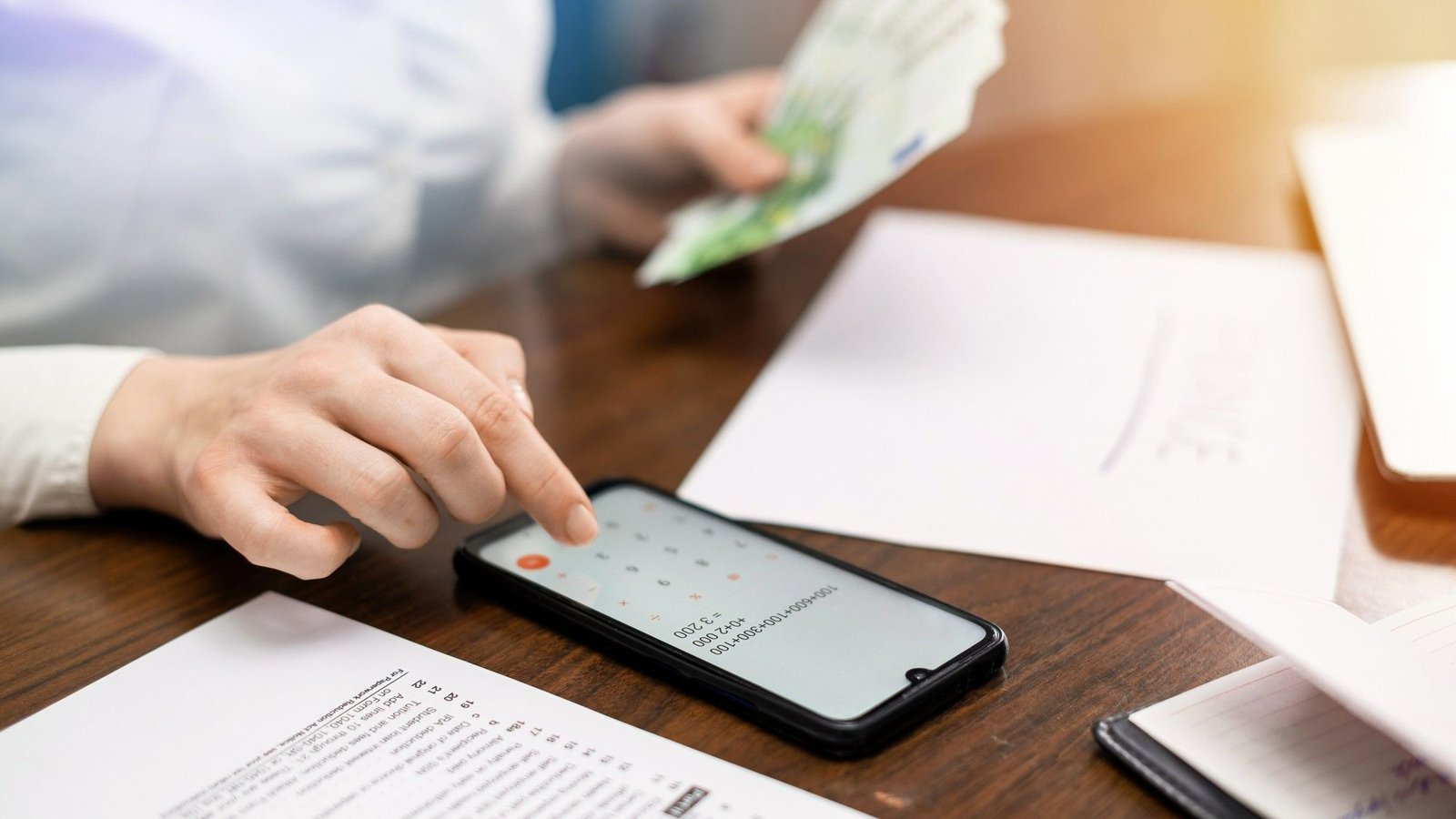 Close-up das mãos de uma mulher usando um smartphone para fazer cálculos financeiros. Ao fundo, ela segura notas de dinheiro sobre uma mesa de madeira com documentos, representando o payback de projetos tecnológicos.
