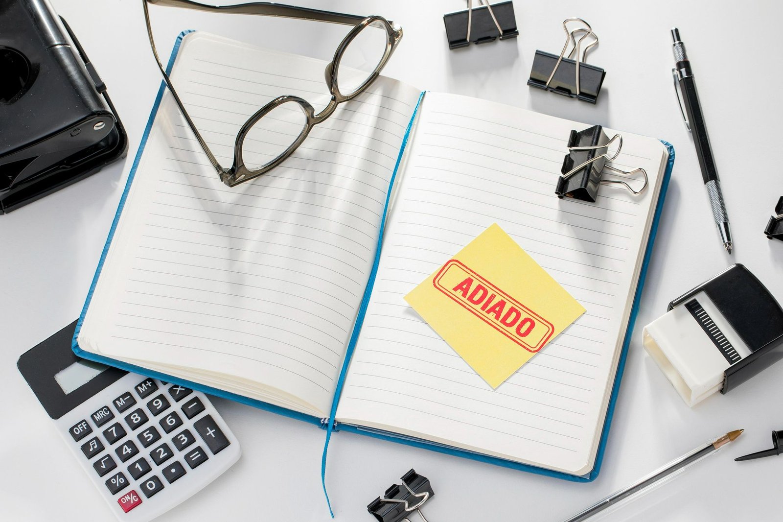 Visão aérea de uma mesa de escritório branca com uma agenda aberta, óculos de grau, calculadora e materiais de papelaria. No centro da agenda, um bilhete amarelo exibe o carimbo vermelho com a palavra "ADIADO". A imagem ilustra a necessidade de organização e políticas de cancelamento claras na gestão de reservas de grupos.