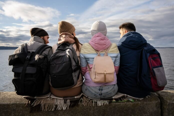 Quatro jovens de costas, com mochilas e casacos, sentados em uma mureta à beira da água no que parece ser uma viagem de lazer. A cena representa o desafio operacional de como gerenciar reservas de grupos com sucesso na hotelaria.