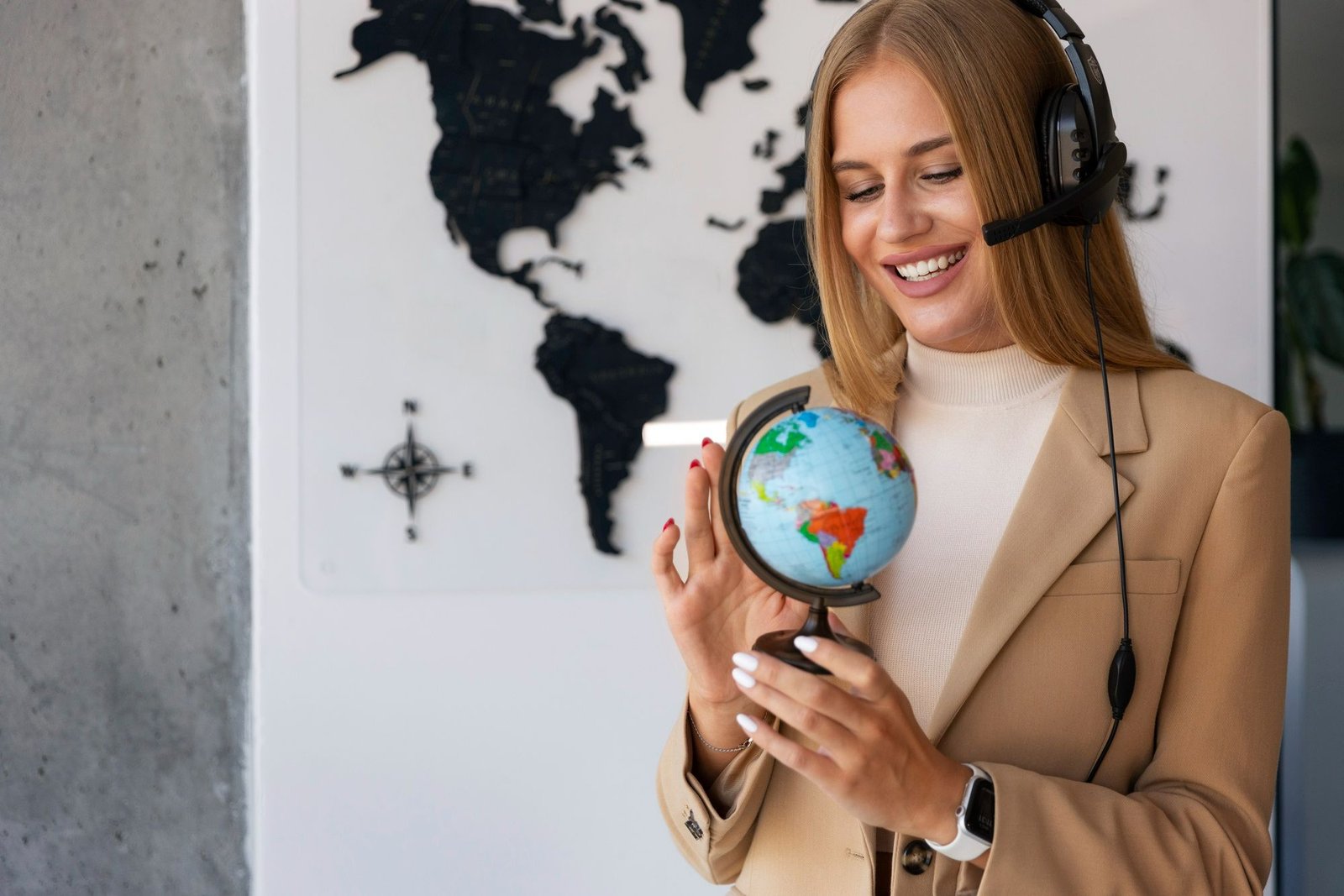 Mulher sorridente com headset e blazer bege manuseia um pequeno globo terrestre, simbolizando o alcance global e a gestão de reservas. Ao fundo, um mapa-múndi e uma rosa dos ventos reforçam a ideia de planejamento e disponibilidade. A imagem ilustra a colaboração entre hotéis e agências na formatação de pacotes seguros e estratégicos.