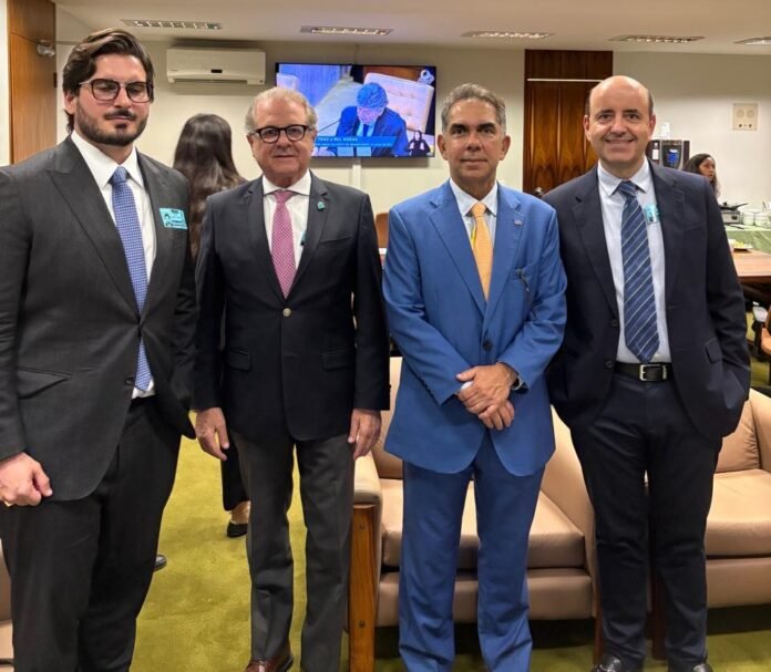 Fotografia em plano médio de quatro homens de terno, em pé e sorrindo, durante encontro que promoveu o debate acerca do fim da escala 6x1. Da esquerda para a direita estão: Leonardo Volpatti, da Volpatti Advogados Associados, vestindo terno cinza-escuro e óculos; Orlando de Souza, do FOHB, com terno preto e gravata cor-de-rosa; Sérgio Gaspar Pereira, da ABIH Nacional, com terno azul-claro; e Antonio Dias, da ABIH-SP, vestindo terno azul-marinho. Ao fundo vê-se um monitor de TV transmitindo uma sessão e parte de um sofá de couro.
