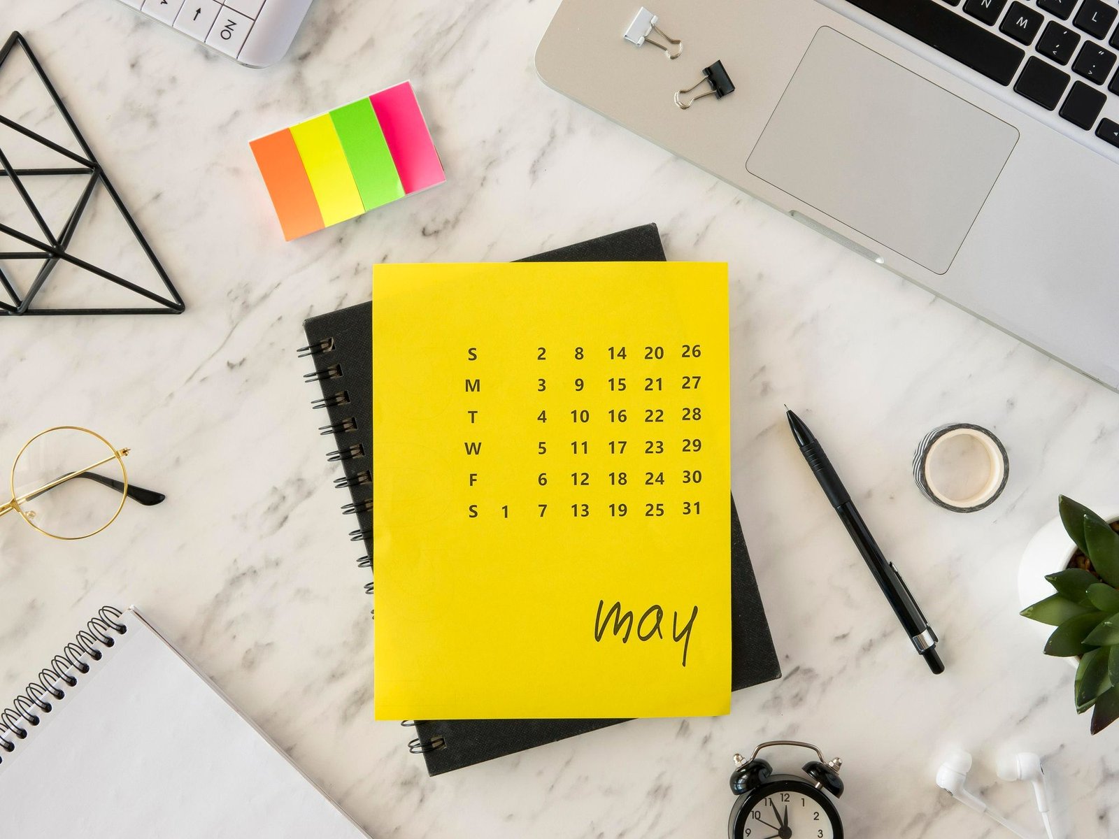 Uma foto em plano superior de uma mesa de escritório organizada, com um notebook, um caderno aberto e, no centro, um calendário amarelo vibrante mostrando o mês de maio. O calendário está destacado e rodeado por itens como uma caneta, óculos de grau, um despertador, fones de ouvido e uma pequena planta.