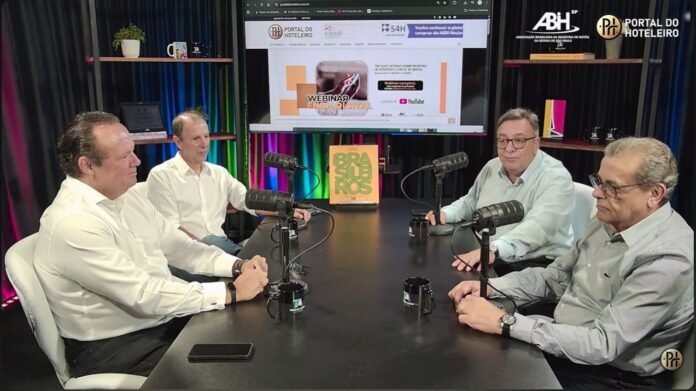 Um registro fotográfico em plano médio de quatro homens sentados ao redor de uma mesa redonda de madeira em um estúdio de gravação para o quadro “Pauta Hoteleira”. O ambiente possui iluminação suave e tons quentes, com microfones profissionais de podcast posicionados em frente a cada participante. No centro da mesa, há uma caneca preta com o logotipo do Portal do Hoteleiro e, ao fundo, um painel com os logotipos da ABIH-SP e do Portal do Hoteleiro. A foto captura o momento de um debate franco e descontraído sobre a FNRH digital, com os participantes interagindo e expressando suas visões sobre o tema.