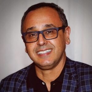 Uma fotografia de estúdio de rosto e ombros de Beto Marques, um homem de meia-idade com cabelo castanho curto e óculos de armação azul, sorrindo amplamente para a câmera. Ele veste um blazer azul-marinho xadrez sobre uma camisa polo preta. O fundo é cinza-claro e desfocado.