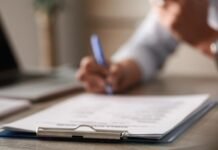 Por que as novas regras trabalhistas correm o risco de impulsionar a informalidade no Brasil Foto em primeiro plano de uma prancheta com papéis sobre uma mesa de escritório, simbolizando a gestão de documentos trabalhistas e o combate à informalidade. Ao fundo, de forma desfocada, uma pessoa segura uma caneta azul, representando o processo de formalização ou contratação de funcionários.