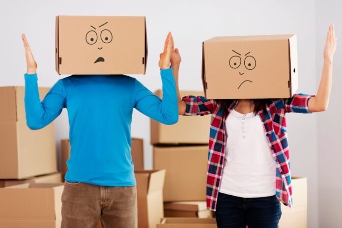 Duas pessoas em pé, lado a lado, com caixas de papelão cobrindo suas cabeças. As caixas possuem desenhos de rostos com expressões de confusão e frustração. A imagem representa o impacto dos erros de estoque na rotina e no clima da equipe hoteleira.