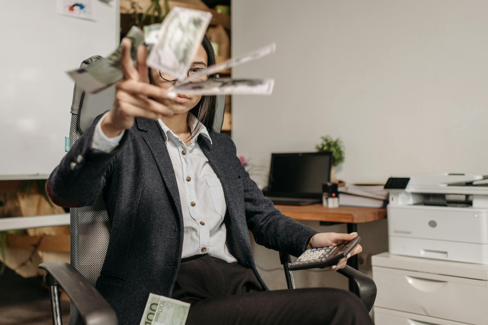 Uma mulher sentada em uma cadeira de escritório, segurando uma calculadora em uma mão e jogando notas de dinheiro para o alto com a outra. O dinheiro aparece desfocado em movimento, simbolizando o desperdício financeiro em um ambiente de trabalho.