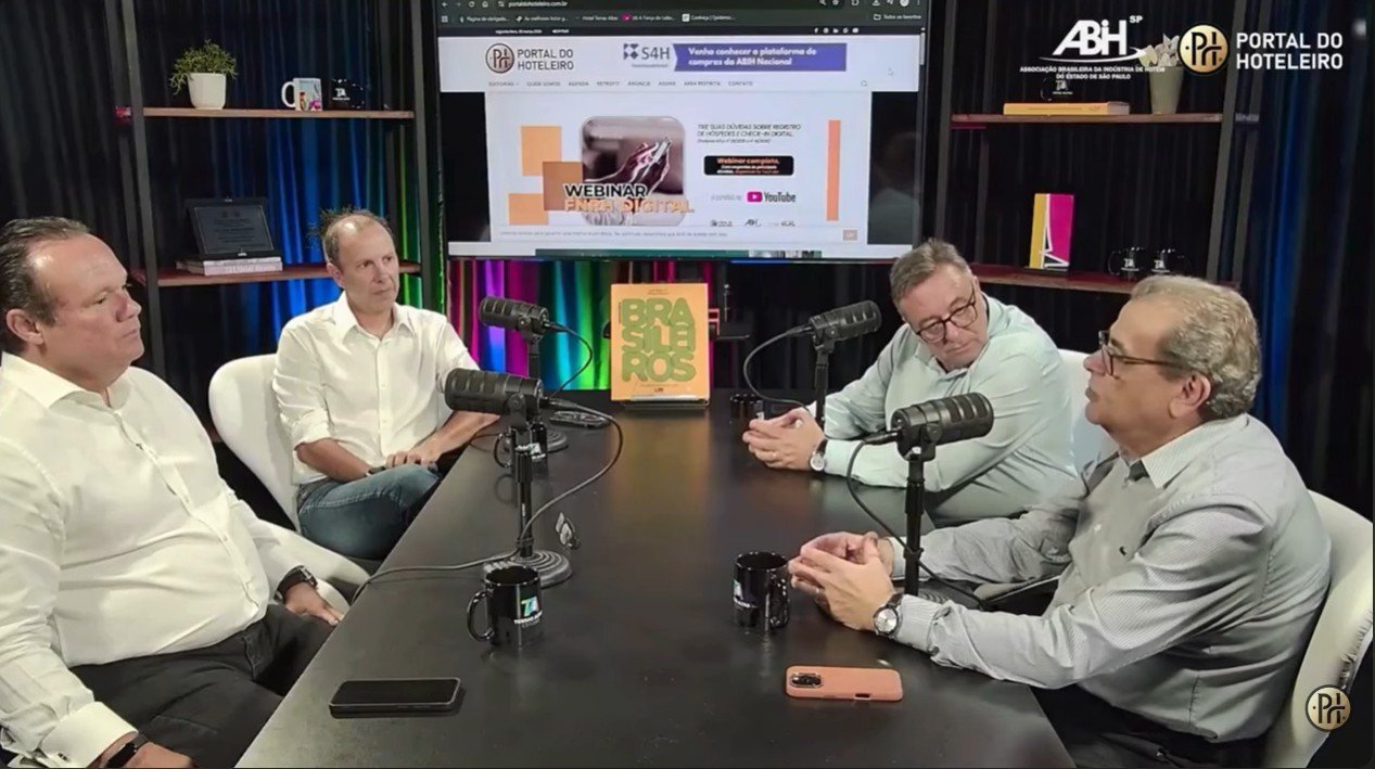 Quatro homens estão sentados ao redor de uma mesa de estúdio preta durante a gravação do videocast do Portal do Hoteleiro sobre a redução da jornada de trabalho na hotelaria. À direita, Luiz Henrique Miranda, gestor do Portal, e Latif Abrão Jr., vice-presidente da ABIH-SP. À esquerda, Marcos Vilas Boas e Gustavo Jarussi, presidente e vice-presidente, respectivamente, da entidade paulista. Todos vestem roupa social e conversam diante de microfones profissionais de rádio. Ao fundo, uma televisão exibe o logotipo do Portal do Hoteleiro e prateleiras com objetos decorativos completam o cenário do estúdio.