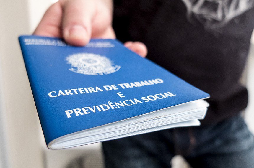 Foto em primeiro plano com foco reduzido, mostrando a mão de uma pessoa estendendo uma Carteira de Trabalho e Previdência Social (CTPS) azul. O documento brasileiro está em destaque na diagonal, sendo possível ler claramente as inscrições em letras douradas na capa. Ao fundo, de forma desfocada, percebe-se o tronco de uma pessoa vestindo uma camiseta escura.