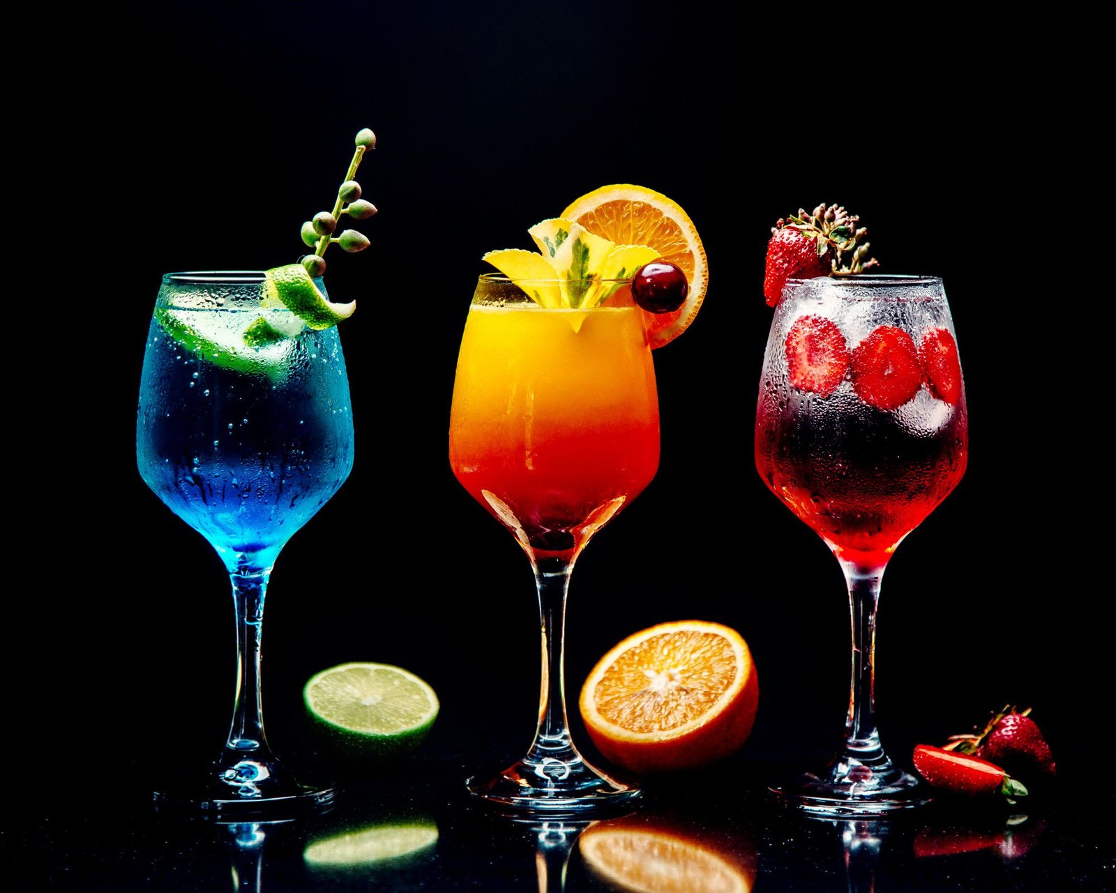 Três mocktails sofisticados servidos em taças de cristal, demonstrando um visual idêntico ao de coquetéis clássicos. À esquerda, uma bebida azul vibrante com guarnição de limão; ao centro, um drink em degradê tropical de amarelo e laranja com frutas frescas; à direita, uma opção refrescante em tons de vermelho com fatias de morango. A composição, sobre fundo preto e superfície reflexiva, destaca o uso de técnicas de mixologia e apresentações artísticas para o público que não consome álcool.