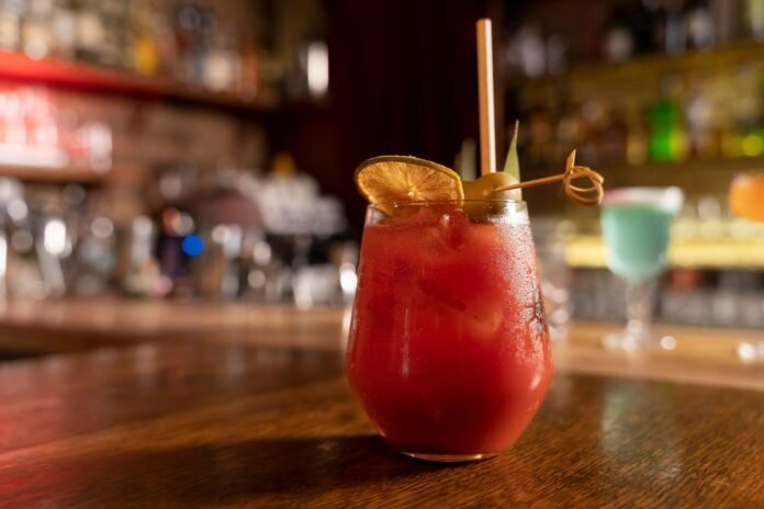 Um mocktail de cor vermelha vibrante servido em um copo baixo de cristal sobre um balcão de madeira. A bebida, que exemplifica a sofisticação das bebidas sem álcool, é decorada com uma fatia de limão siciliano seco, azeitonas em um palito artesanal e um canudo sustentável, com o ambiente do bar ao fundo desfocado.