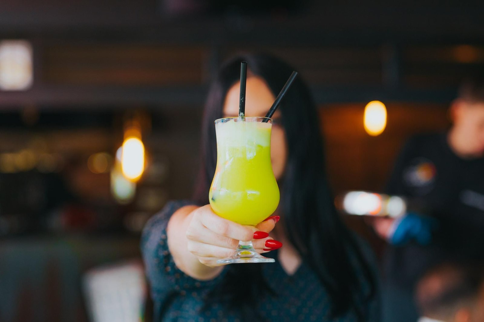 Uma jovem com unhas pintadas de vermelho segura uma taça de mocktail em tons de verde cítrico com espuma densa, estendendo-a em um gesto de brinde. A imagem captura a essência da nova tendência de bebidas sem álcool, na qual a apresentação moderna e o ambiente sofisticado do bar elevam a experiência de consumo para além das opções tradicionais.