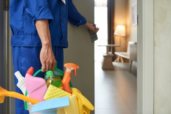 Em close, funcionário vestindo uniforme azul segura um balde repleto de produtos de limpeza coloridos, esponjas e luvas amarelas, enquanto abre a porta de um quarto de hotel iluminado ao fundo. O uso de insumos específicos e adequados reforça a importância da limpeza profissional para a hotelaria, garantindo a higiene e o bem-estar dos hóspedes.