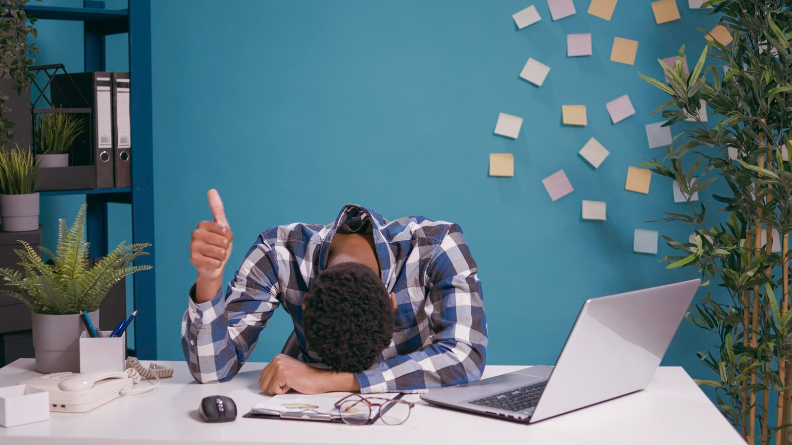 Homem com a cabeça debruçada sobre a mesa de trabalho, demonstrando exaustão, enquanto faz um sinal de "joinha" com a mão. O cenário ilustra o desgaste na rotina da recepção de hotel, com um notebook aberto, diversos post-its espalhados na parede azul ao fundo e papéis acumulados, simbolizando a sobrecarga e a desorganização operacional.