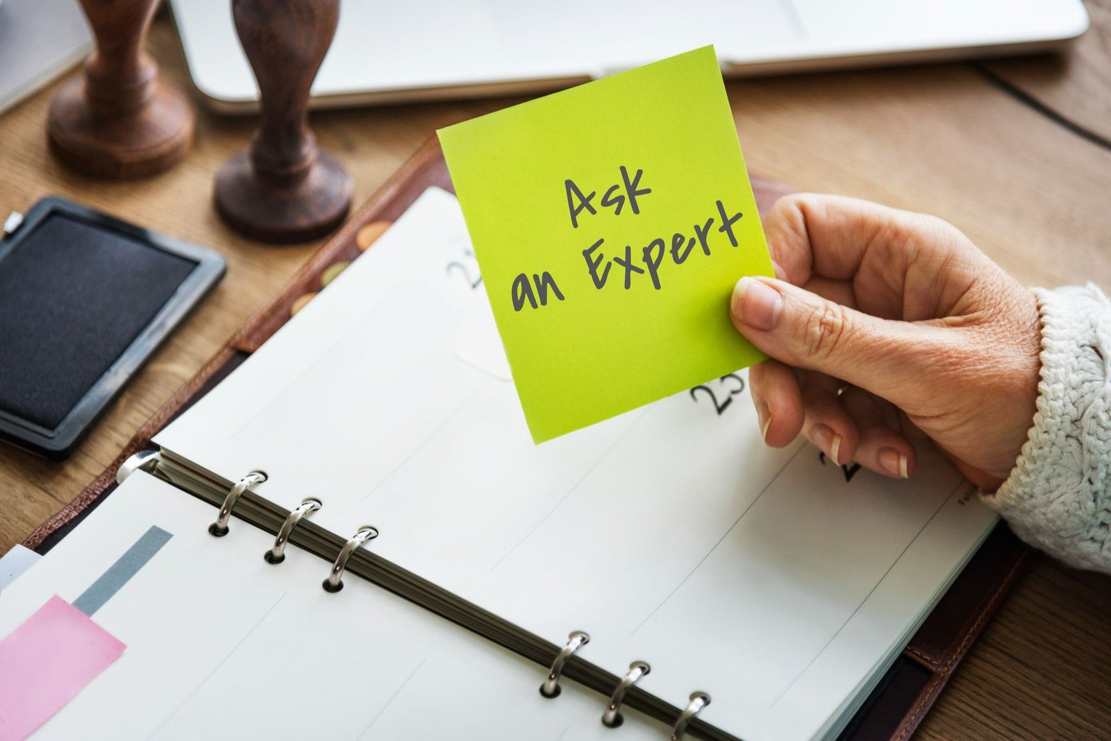 Uma mão segura um post-it verde com a frase "Ask an expert" ("pergunte a um especialista") sobre uma agenda aberta em uma mesa de madeira. A cena representa a interrupção na rotina da recepção de hotel quando o profissional, sem autonomia para decidir, precisa paralisar o atendimento para buscar validação da supervisão.