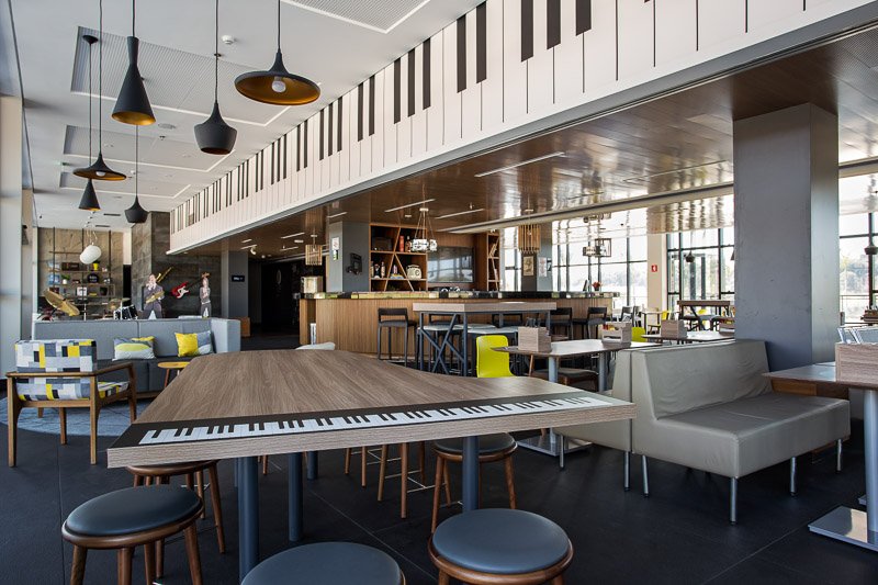 Área social do hotel Ibis Styles Taubaté, com decoração temática musical. No primeiro plano, uma mesa de madeira em formato de piano de cauda com a estampa de um teclado na borda. Ao fundo, uma sanca no teto também reproduz teclas de piano em grande escala. O ambiente moderno conta com pendentes pretos e dourados, sofás em tons de cinza com almofadas amarelas, cadeiras de design contemporâneo e um bar ao fundo com prateleiras de madeira. O espaço é amplo, iluminado por grandes janelas, reforçando o conceito lúdico e instagramável do projeto da ARTEO.