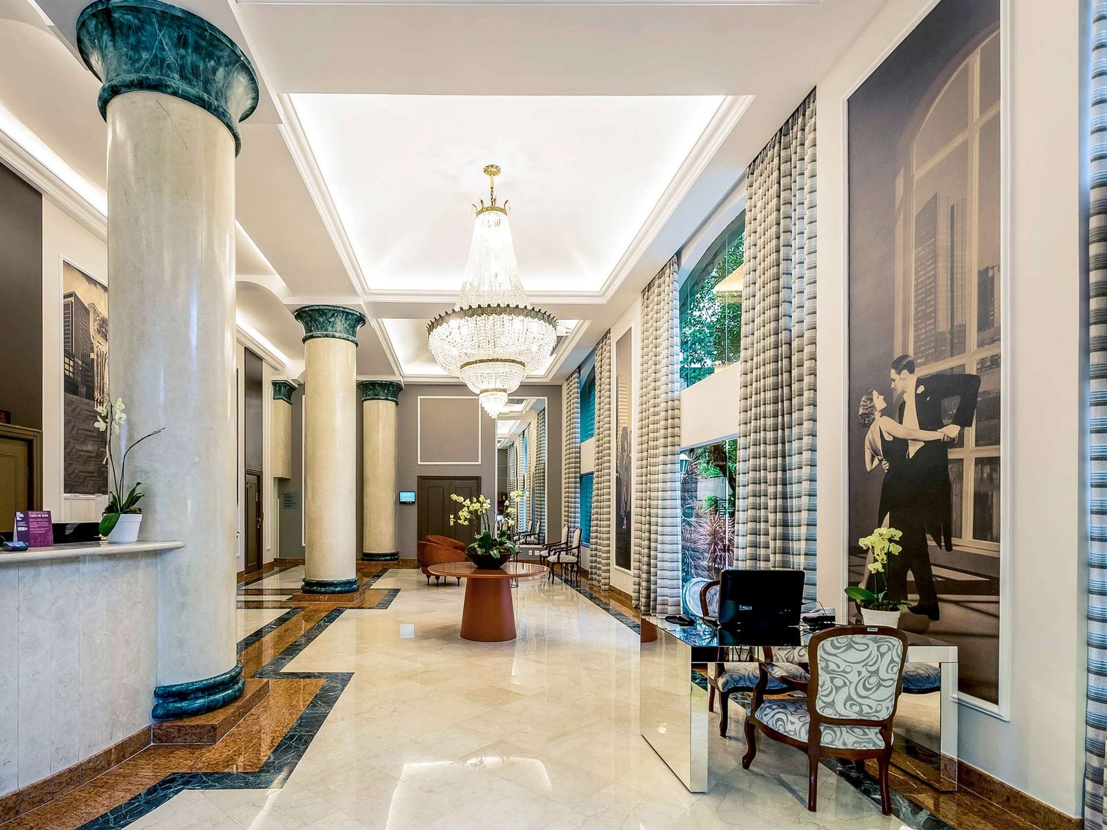 Fotografia da recepção do hotel Mercure São Paulo JK, exibindo um design clássico e elegante. O lobby possui pé-direito alto com grandes colunas de mármore claro e capitéis em tom verde-esmeralda. Ao centro, destaca-se um suntuoso lustre de cristal clássico. Nas paredes, painéis artísticos em preto e branco retratam cenas urbanas e casais em trajes de gala, conectando o hotel à história do bairro. O ambiente conta com piso de mármore polido, cortinas longas em tons neutros e um balcão de recepção em espelho, unindo tradição e modernidade no projeto assinado pela ARTEO.