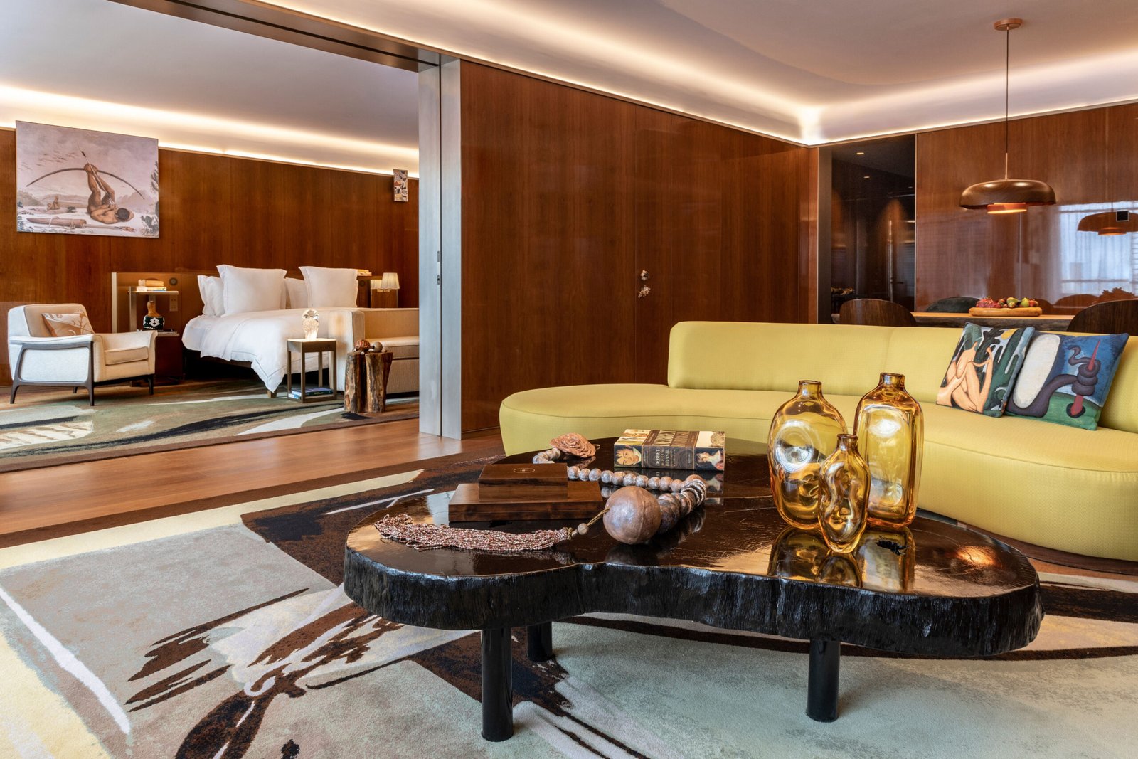 Suíte de luxo no Rosewood São Paulo, exemplificando o design iconográfico. O ambiente possui paredes revestidas em madeira de tom quente e iluminação indireta no teto. Em destaque, uma sala de estar com sofá curvo amarelo, mesa de centro orgânica em madeira escura com vasos de vidro âmbar e um tapete amplo com padrões artísticos. Ao fundo, através de uma abertura na parede, vê-se o quarto com cama branca, poltrona clara e uma tela na parede retratando uma figura indígena, reforçando a curadoria de arte brasileira do projeto.