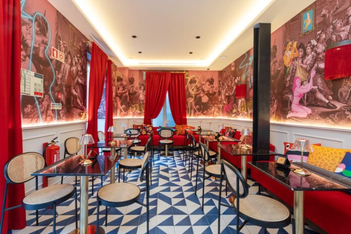 Fotografia de interiores mostrando o bar do Upon Angels Lisboa. O ambiente tem paredes revestidas por papel de parede de gravuras clássicas misturadas com elementos pop, como seções destacadas e luzes neon. O chão é um mosaico geométrico azul e branco. O mobiliário inclui mesas de mármore preto com luminárias de mesa em acrílico, cadeiras de palhinha e bancos em veludo vermelho, que combinam com as cortinas teatrais vermelhas. O projeto de design iconográfico da ARTEO criou um espaço lúdico e sofisticado.