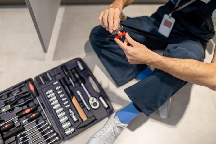 Sentado no chão, um profissional de manutenção segura uma pequena peça hidráulica com válvula vermelha, enquanto uma maleta de ferramentas aberta repousa ao seu lado. A imagem detalha o cuidado técnico necessário na manutenção em hotéis, destacando a organização e o preparo da equipe para reparos preventivos e corretivos.