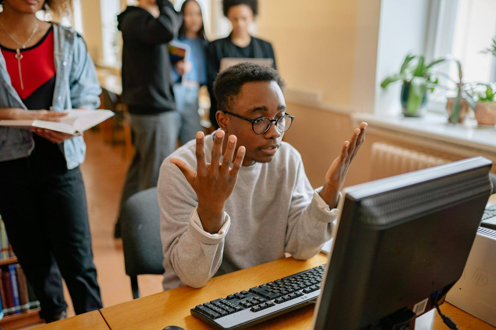 Um jovem homem negro, usando óculos e um moletom cinza, está sentado em uma mesa de madeira, olhando para a tela de um computador desktop com uma expressão de total confusão e frustração, erguendo as duas mãos abertas em um gesto de desabafo. Ao fundo, outras pessoas desfocadas circulam, uma delas segurando um caderno. A cena representa o momento em que a equipe de um hotel se sente "refém" de um sistema complexo e engessado, dificultando a eficiência operacional e o atendimento fluido ao hóspede.