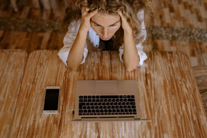 Mulher jovem com cabelos cacheados, sentada à mesa de madeira, olha para a tela de um notebook com expressão de cansaço e frustração, apoiando a cabeça nas mãos. Sobre a mesa, há também um smartphone ao lado do computador. A cena ilustra a complexidade e as eventuais dificuldades envolvendo o uso de tecnologia na hotelaria, especialmente quando não há um planejamento por trás da mudança.