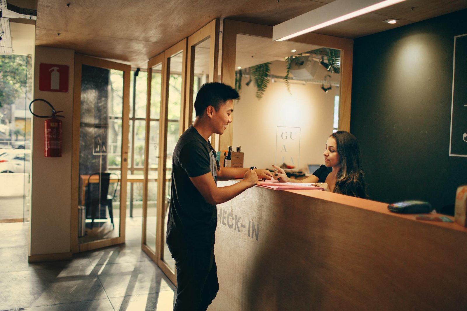 Recepcionista de hotel atende um hóspede no balcão de check-in em um ambiente moderno com iluminação acolhedora. Ambos sorriem enquanto interagem sobre o balcão de madeira, sobre o qual se vê uma máquina de cartão e documentos. A cena exemplifica o equilíbrio ideal no uso de tecnologia na hotelaria: a ferramenta digital agiliza o processo, mas o foco principal permanece na hospitalidade e na conexão humana entre a equipe e o cliente.