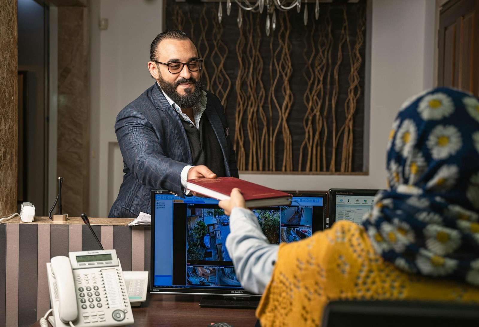 Uma recepcionista de hotel, usando um hijab floral e colete amarelo, entrega um livro ou passaporte a um hóspede no balcão de atendimento. À frente dela, sobre o balcão, há um telefone e monitores que exibem o sistema de câmeras de segurança do estabelecimento. A imagem retrata o suporte da tecnologia na hotelaria para o controle e segurança, enquanto a colaboradora mantém o foco no atendimento direto e humano ao cliente.