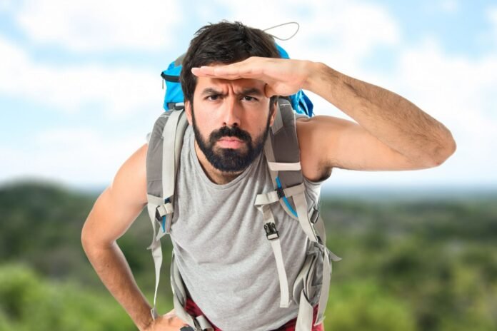 Fotografia de um homem jovem com barba e mochila de trilha nas costas, inclinando o corpo para frente e fazendo uma 
