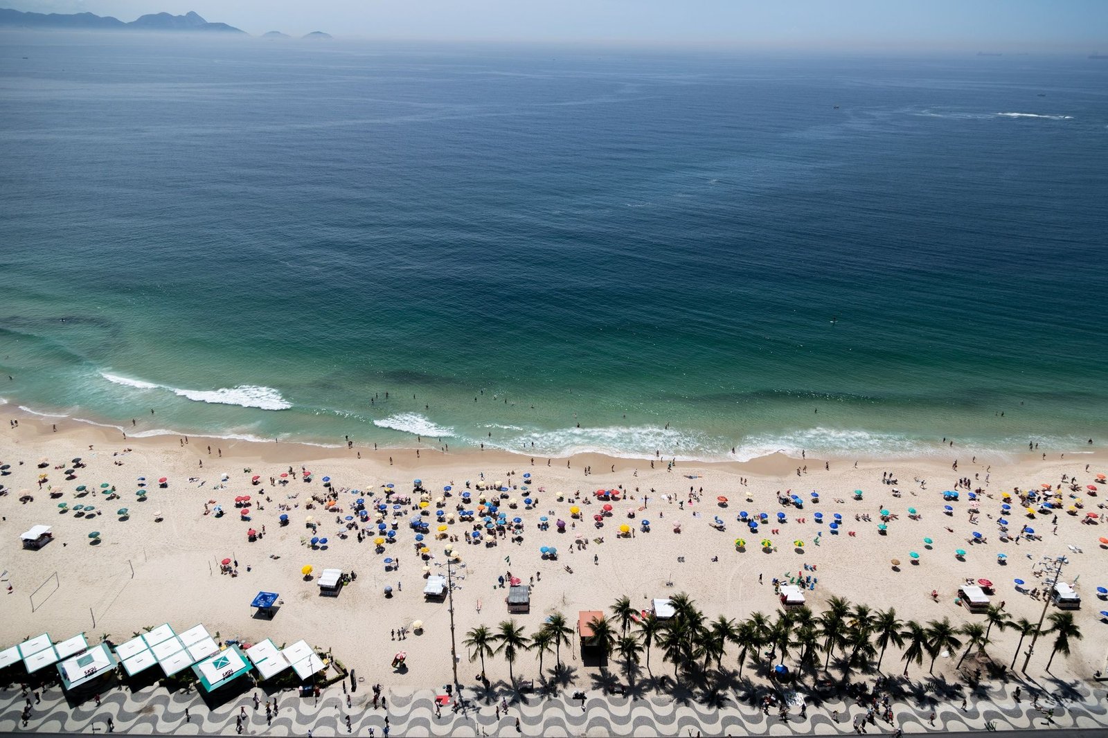 Fotografia aérea da orla de uma praia movimentada. A imagem mostra o contraste entre o azul intenso do mar, a areia clara repleta de guarda-sóis coloridos e o calçadão com desenhos ondulados em preto e branco, ladeado por coqueiros. O cenário ilustra a alta temporada e a preferência dos viajantes por destinos de verão.