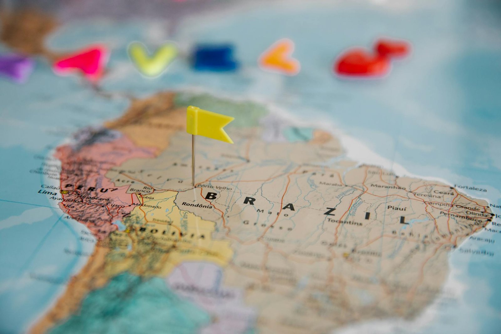 Fotografia de um mapa da América do Sul com foco no Brasil. Um marcador amarelo em formato de bandeira está fincado sobre o mapa, simbolizando a escolha de um destino. Ao fundo, letras coloridas aparecem desfocadas. A imagem ilustra o planejamento de viagens nacionais e o desafio de como atrair hóspedes para destinos brasileiros.