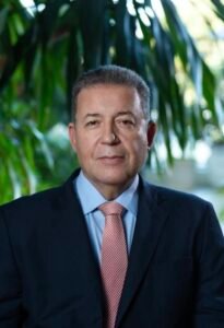 Foto de estilo retrato de Alexandre Sampaio, da FBHA. Homem de cabelos curtos grisalhos, olhando para a frente com expressão séria. Ele veste paletó preto, camisa social azul-clara e gravata com padronagem avermelhada, posando com folhas verdes de plantas ao fundo.
