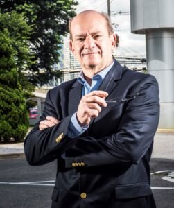 Foto de estilo retrato de João de Nagy. Homem calvo de cabelos curtos nas laterais, olhando para a frente com os braços cruzados e segurando um par de óculos na mão esquerda. Ele veste um blazer azul-marinho com botões dourados sobre camisa social azul-clara, posando em um ambiente externo com árvores e uma estrutura urbana ao fundo.