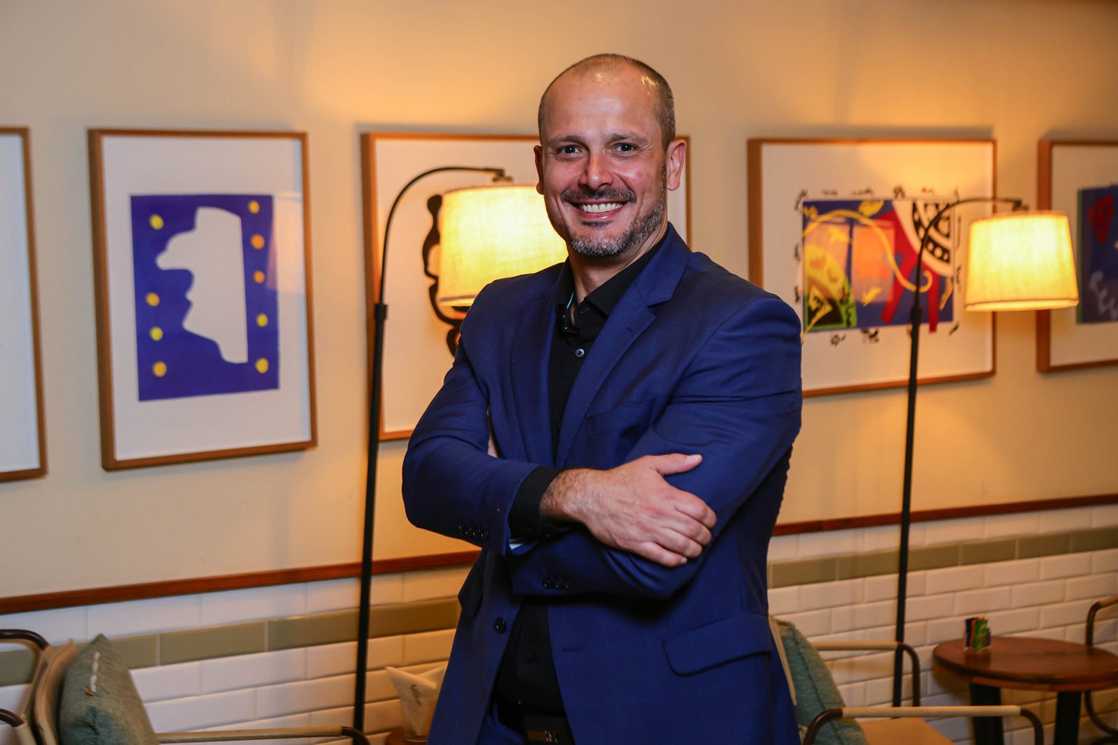 Foto de meio corpo de Thiago Borges, da Resorts Brasil. Homem de cabelo raspado e barba curta, sorrindo com os braços cruzados. Ele veste paletó azul sobre camisa preta, posando em um ambiente interno decorado com quadros e luminárias ao fundo.