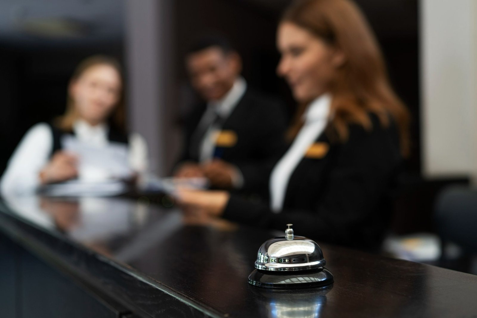 Uma sineta metálica de recepção em destaque sobre um balcão escuro e brilhante. Ao fundo e fora de foco, três funcionários uniformizados trabalham atrás do balcão do front desk, representando o envolvimento da equipe de recepção nas estratégias de vendas do hotel.